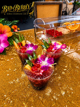 Three festive tropical champagne cocktails with crushed ice, maraschino cherries, pineapple wedges and purple orchid garnishes on a gold glitter event table with a citrus station in the background.