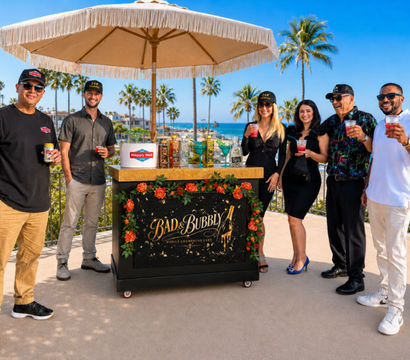 Six people holding colorful cocktails around a floral-decorated mobile bar under a fringed umbrella on an oceanfront patio with palm trees and bright blue sky — coastal resort vibe.
