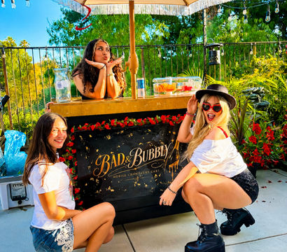 Three friends posing at a decorated outdoor drink cart under a large umbrella on a sunny backyard patio, surrounded by string lights, lush greenery and festive summer brunch vibes.