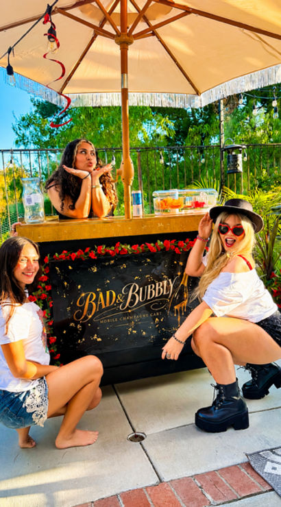 Three friends posing at a decorated outdoor drink cart under a large umbrella on a sunny backyard patio, surrounded by string lights, lush greenery and festive summer brunch vibes.