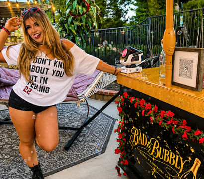 Smiling woman striking a playful pose on a festive backyard patio beside a floral-decorated mobile cocktail cart under a fringed umbrella, hammock, string lights and trees at dusk