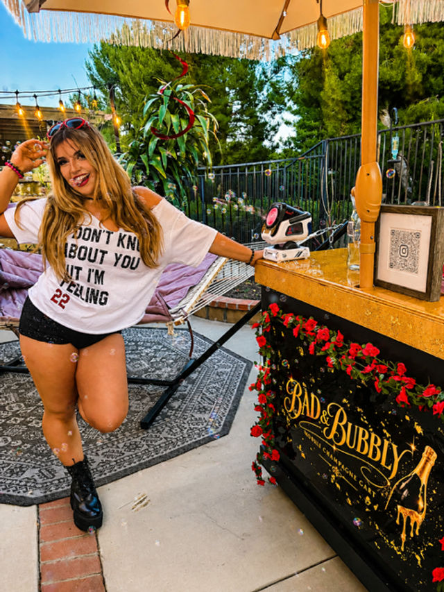 Smiling woman striking a playful pose on a festive backyard patio beside a floral-decorated mobile cocktail cart under a fringed umbrella, hammock, string lights and trees at dusk