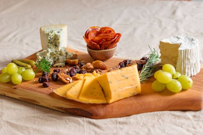 Gourmet charcuterie board on a wooden serving board with herb-speckled cheddar wedges, creamy brie, blue cheese crumbles, a cup of pepperoni, green grapes, cornichons, mixed nuts and dried fruit, garnished with rosemary.
