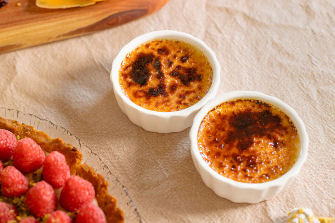 Close-up of two classic French crème brûlées in white ramekins with golden crackled sugar tops on a linen tablecloth, a raspberry tart partially visible at the edge.