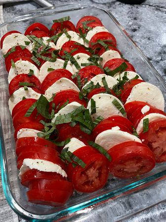 Bright Caprese salad with alternating slices of ripe red tomato and fresh mozzarella, topped with sliced basil and cracked black pepper in a glass dish