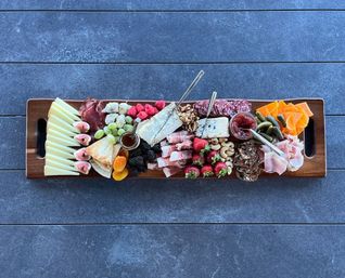 Long wooden charcuterie board on a dark stone tabletop with assorted cheeses and brie, cured meats, grapes, figs, strawberries and blackberries, dried apricots, nuts, crackers, cornichons, and small jars of honey and jam — colorful and ready to share.