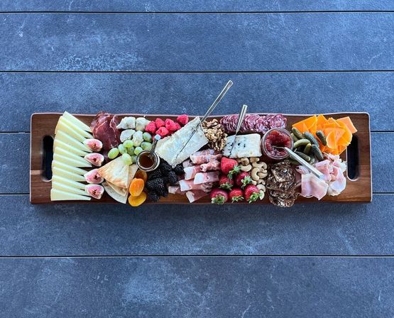 Long wooden charcuterie board on a dark stone tabletop with assorted cheeses and brie, cured meats, grapes, figs, strawberries and blackberries, dried apricots, nuts, crackers, cornichons, and small jars of honey and jam — colorful and ready to share.