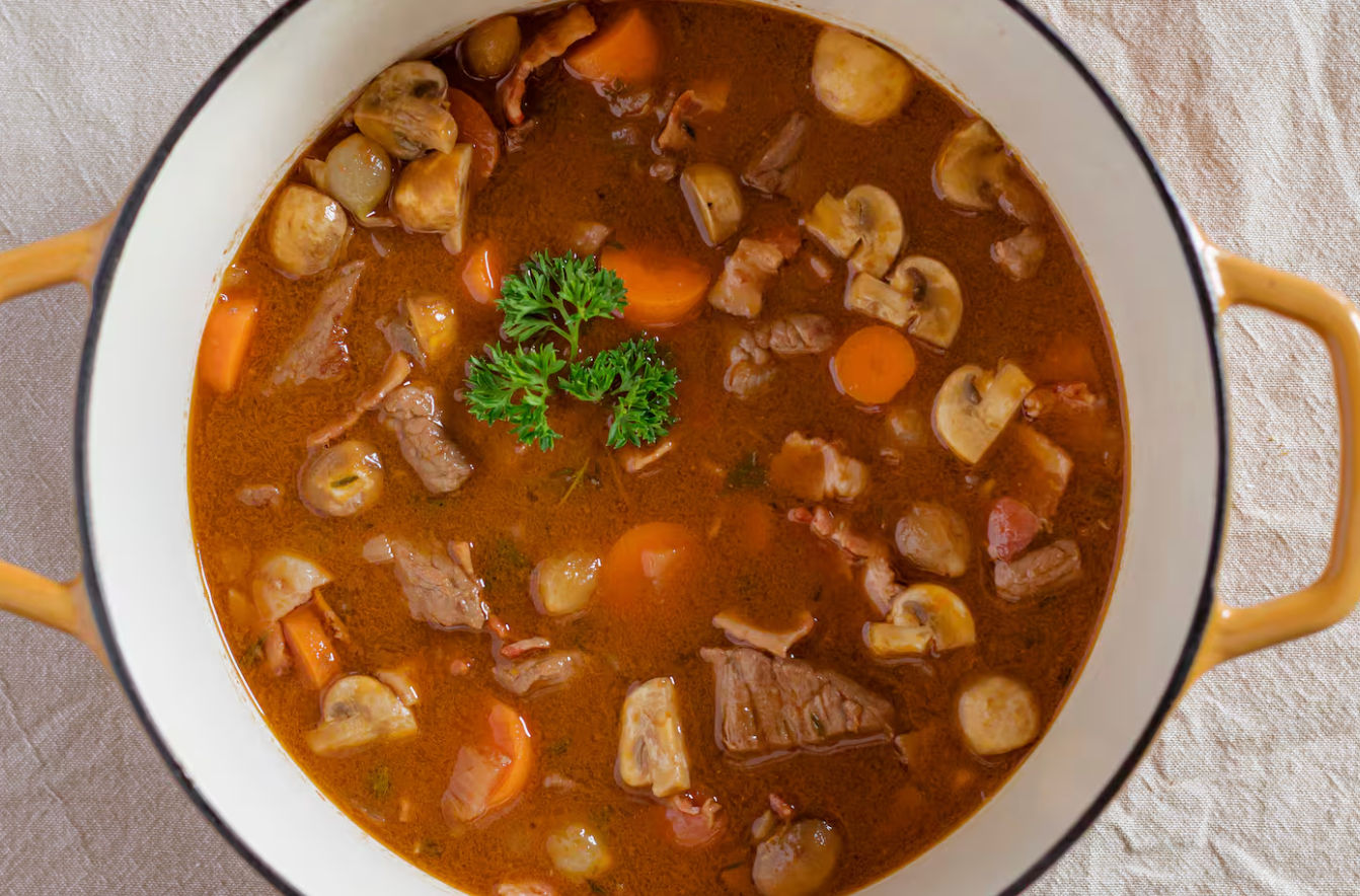 Top-down view of a hearty beef and mushroom stew with sliced mushrooms, carrot rounds, pearl onions and parsley garnish in an enameled pot with yellow handles on a linen cloth
