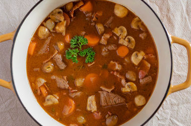 Top-down view of a hearty beef and mushroom stew with sliced mushrooms, carrot rounds, pearl onions and parsley garnish in an enameled pot with yellow handles on a linen cloth