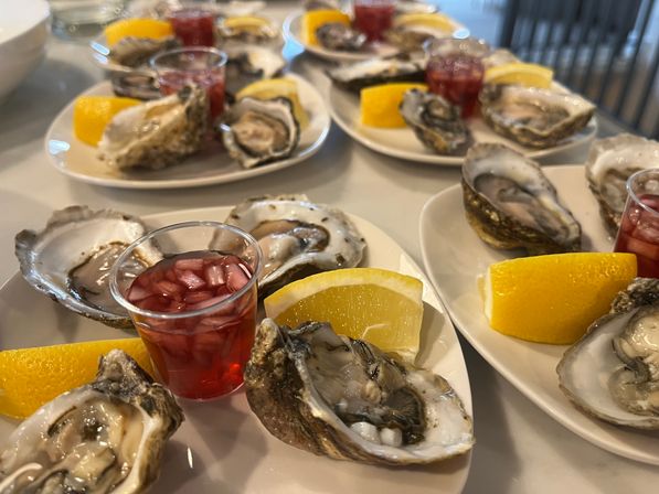 Close-up of fresh shucked raw oysters on half shells arranged on white plates with lemon wedges and small cups of red mignonette sauce, seafood appetizer spread