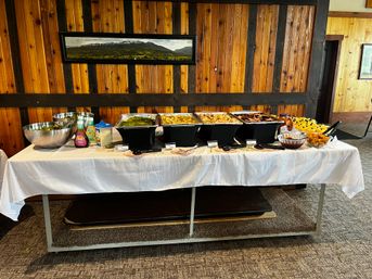 Savory buffet spread on a white-clothed table in a rustic wood-paneled banquet room — chafing dishes with veggies, potatoes, meats, salad bowls, condiments and desserts beneath a mountain panorama painting.