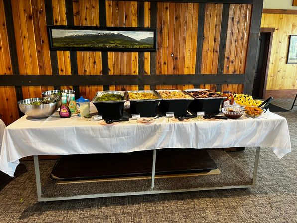 Savory buffet spread on a white-clothed table in a rustic wood-paneled banquet room — chafing dishes with veggies, potatoes, meats, salad bowls, condiments and desserts beneath a mountain panorama painting.
