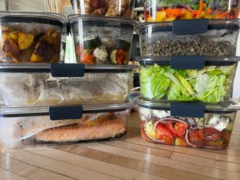 Stacked glass meal-prep containers on a wooden kitchen counter with a salmon fillet, romaine salad, lentils, roasted vegetables and a tomato‑feta salad — colorful healthy meal prep.