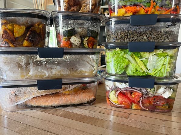 Stacked glass meal-prep containers on a wooden kitchen counter with a salmon fillet, romaine salad, lentils, roasted vegetables and a tomato‑feta salad — colorful healthy meal prep.