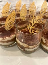 Mini tiramisu cups in clear glasses - layered mascarpone and espresso-soaked cake with cocoa dusting and leaf-shaped white chocolate toppers, arranged on a dessert table.