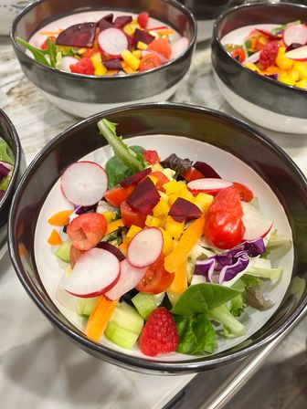 Close-up colorful mixed salad bowl with radish slices, cherry tomatoes, raspberries, diced beet and yellow bell pepper, carrot, cucumber, purple cabbage and mixed greens in a black-and-white ceramic bowl on a marble countertop
