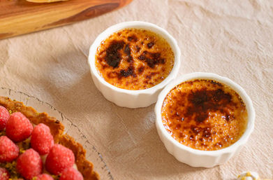Two golden, caramelized crème brûlées in white ramekins on a beige linen tablecloth, with the edge of a fresh raspberry tart visible