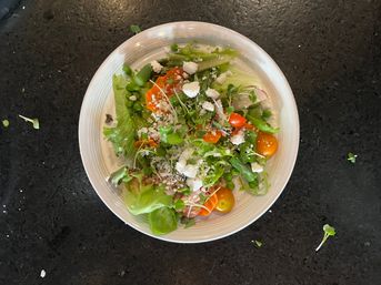 Top-down view of a vibrant mixed-greens salad in a white bowl with cherry tomatoes, peas, microgreens and crumbled feta on a dark countertop.
