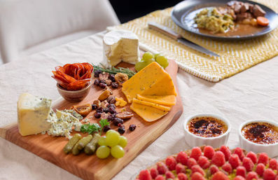 Inviting wooden cheese board with brie, blue cheese, herbed cheddar slices, folded salami, cornichons, green grapes, dried figs, nuts and dried cranberries on a linen table, with two crème brûlée cups and a raspberry tart nearby.