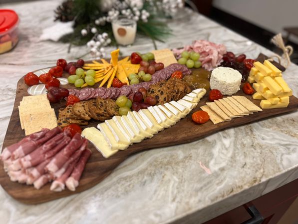 Wooden charcuterie board on a marble countertop with assorted cheeses (brie wedges, cubed cheddar, herbed goat cheese), sliced salami and prosciutto, crackers and fruit crisps, green and red grapes, roasted cherry tomatoes and jam — festive appetizer spread.