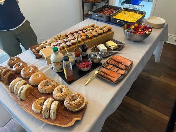 Home-style brunch buffet on a white tablecloth: assorted bagels on wooden boards with cream cheese, butter and jams, smoked salmon slices on a platter, mini muffins, scrambled eggs and bacon in pans, and a bowl of mixed fresh fruit.