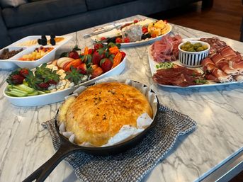 Golden baked brie in a cast-iron skillet on a marble countertop with charcuterie platters of prosciutto, salami, cheeses, olives, fresh strawberries and crackers — cozy indoor appetizer spread.