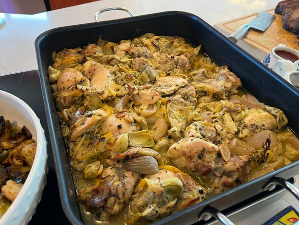 Roasted herb chicken thighs with artichoke hearts, caramelized shallots and lemon slices in a large black roasting pan on a kitchen countertop