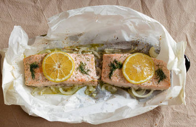 Two oven-baked salmon fillets en papillote on crinkled parchment paper, each topped with a lemon slice and fresh dill, resting on sliced onions