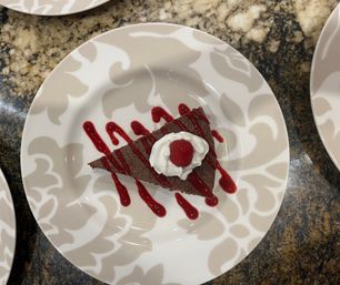 Decadent triangular slice of chocolate cake drizzled with raspberry sauce, topped with whipped cream and a fresh raspberry on a patterned white plate over a granite countertop