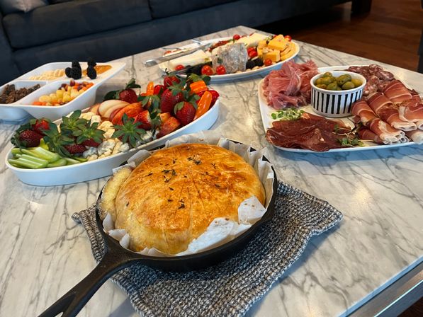 Golden baked brie en croute in a cast-iron skillet on a marble coffee table, surrounded by charcuterie and fruit platters with prosciutto, olives, cheeses, strawberries and crackers — cozy indoor appetizer spread.