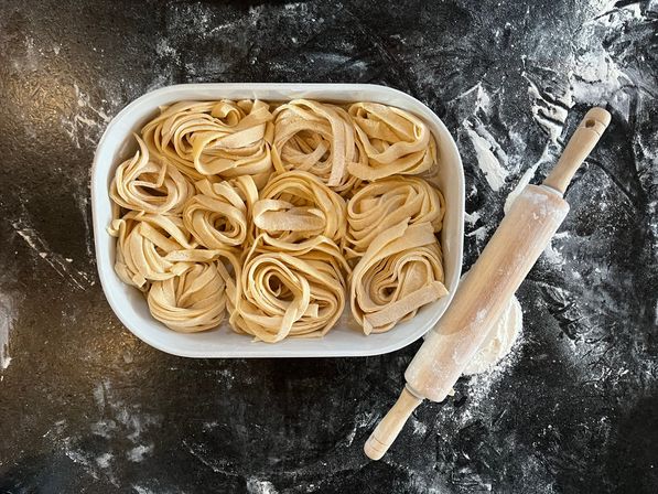 Top-down view of coiled fresh pasta nests in a white oval dish beside a flour-dusted wooden rolling pin on a dark, floured countertop.