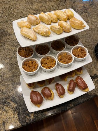 Three-tier white dessert stand on a granite countertop with glazed mini turnovers on top, individual apple-crumble ramekins in the middle, and chocolate-glazed éclairs on the bottom — tempting party dessert display.