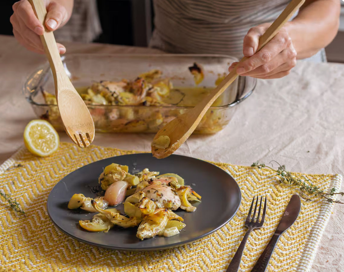 Hands using wooden utensils to serve roasted chicken and vegetables from a glass baking dish onto a gray plate on a yellow patterned placemat with a lemon half and cutlery — cozy home-cooked dinner.