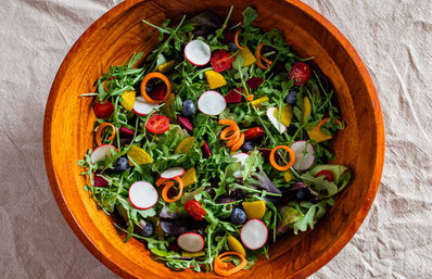 Vibrant mixed greens salad in a wooden bowl on a linen cloth, with arugula, radish slices, cherry tomatoes, carrot curls, golden beets and blueberries