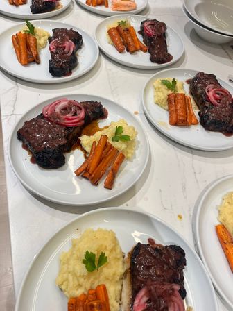 Multiple white plates of steak topped with glossy red-wine sauce and pickled red onions, creamy mashed potatoes and roasted carrots arranged on a white marble countertop ready for dinner service.