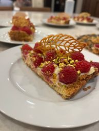 Close-up of a raspberry and pistachio tart slice with golden crumb crust and decorative caramel lattice tuile, served on a white plate at a café-style setting.