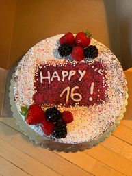 Round berry-topped birthday cake dusted with powdered sugar and red crumbs, piped in white icing to read 'HAPPY 16!', decorated with strawberries, raspberries and blackberries.