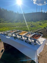 Sunlit outdoor buffet in an alpine meadow — chafing trays of food on a white-clothed table with pine trees, rolling hills and a bright sun flare under a blue sky.