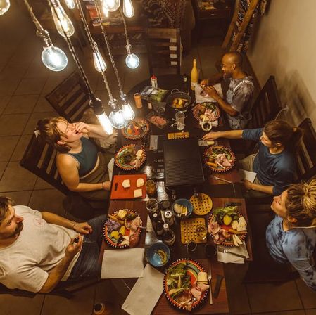 Overhead shot of five people at a cozy home dinner party around a wooden table under hanging Edison bulbs, colorful plates of charcuterie and veggies, a tabletop grill in the center with wine bottles and condiments — casual indoor dining scene.