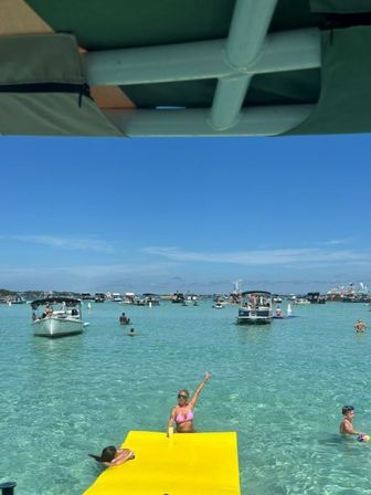 Sunny sandbar scene with clear turquoise water, anchored boats and swimmers, a bright yellow floating mat and a woman in a pink bikini waving under a blue sky.
