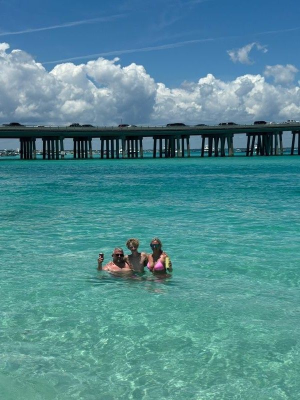 Three people waist-deep in clear turquoise ocean water, two adults and a child smiling with drinks near a long coastal bridge and puffy white clouds on a sunny tropical day.