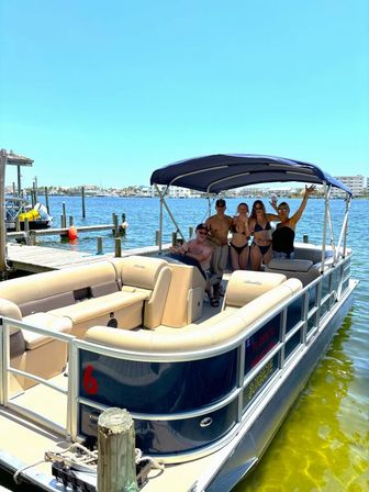 Pontoon boat docked at a sunny marina with a group of adults in swimwear waving under a navy bimini, clear blue sky and calm coastal waters