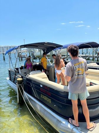 Pontoon boat tied at a sunny coastal marina dock, people boarding in swimwear and summer clothes with waterfront condos and clear blue sky in the background.