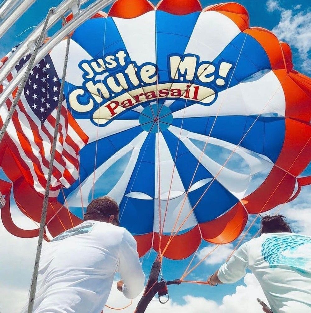 Red, white and blue parasail canopy with an American flag, two crew members securing the lines under a bright blue sky — playful parasailing scene.