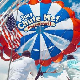 Red, white and blue parasail canopy with an American flag, two crew members securing the lines under a bright blue sky — playful parasailing scene.