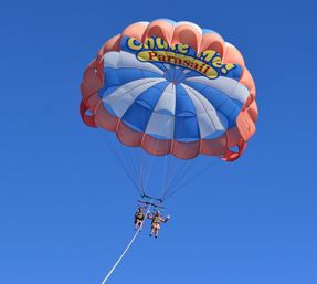 Tandem parasailing ride with two riders suspended beneath a colorful orange-and-blue parachute against a clear blue sky