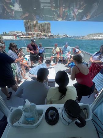 Sunlit harbor boat with passengers gathered on deck around a game or demonstration, marina docks and beachfront high‑rise skyline along the coastline in the background.