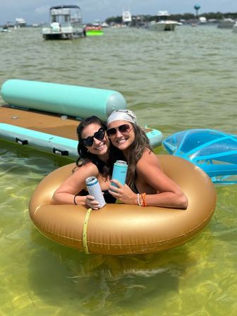 Two friends in sunglasses sharing a gold donut float in shallow green bay water, holding canned drinks with pontoons and boats in the background on a sunny summer boating day