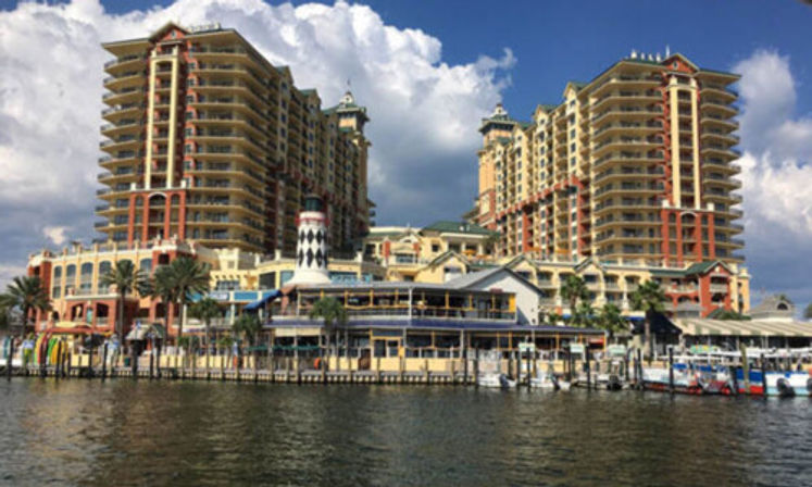 Waterfront resort and marina with twin high-rise towers, a lighthouse-style tower, palm trees and docked boats under a partly cloudy sky.