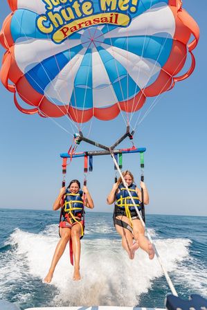 Two riders tandem parasailing over a blue ocean, wearing life jackets and suspended from a colorful red, white and blue canopy above a boat wake under a clear sky.